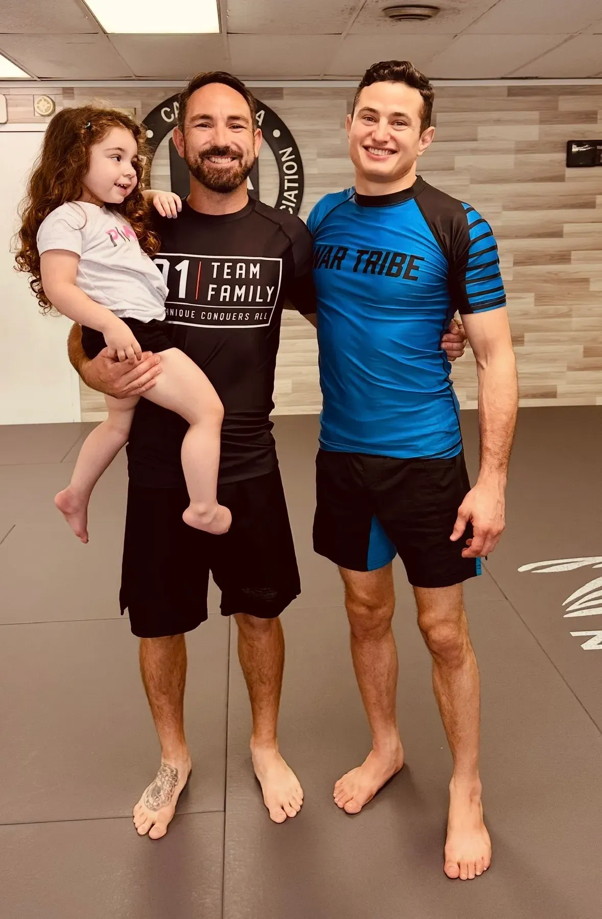 Two adult students and a child stand together on the academy mats after class.